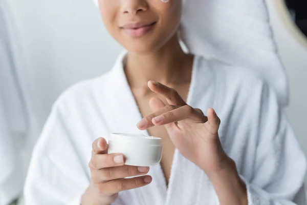 Cropped view of african american woman in bathrobe holding container with face cream — Stock Photo