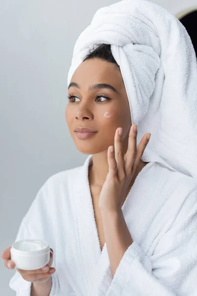 Dreamy african american woman in bathrobe holding container with face cream in bathroom — Stock Photo