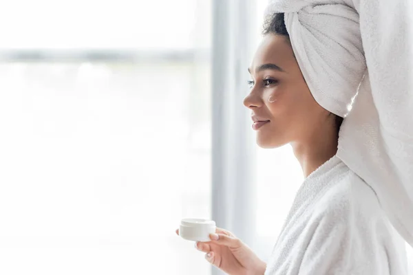 Sonriente mujer afroamericana en albornoz sosteniendo contenedor con crema cosmética en el baño - foto de stock