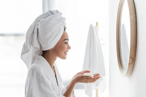 Side view of happy african american woman in bathrobe holding container with face cream and looking at mirror — Stock Photo