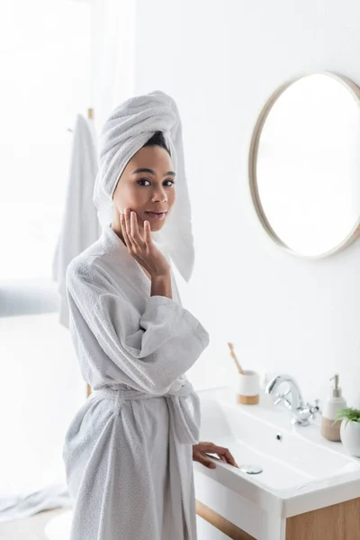 Mujer joven afroamericana en albornoz y toalla aplicando crema facial en el baño - foto de stock