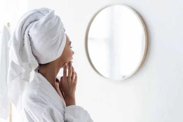 Афроамериканська жінка в халаті, застосовує крем для обличчя і дивиться на дзеркало у ванній кімнаті. — стокове фото