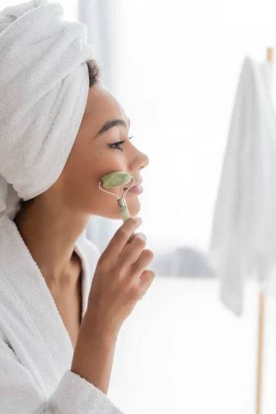 Side view of pleased african american woman in bathrobe and towel massaging face with jade roller in bathroom — Stock Photo