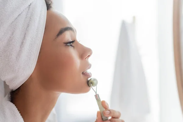 Side view of pretty african american woman in towel massaging face with jade roller in bathroom — Stock Photo