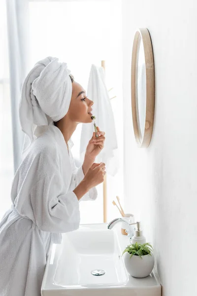 Vista lateral de mujer afroamericana en albornoz y toalla masajeando la cara con rodillo de jade mientras mira el espejo - foto de stock
