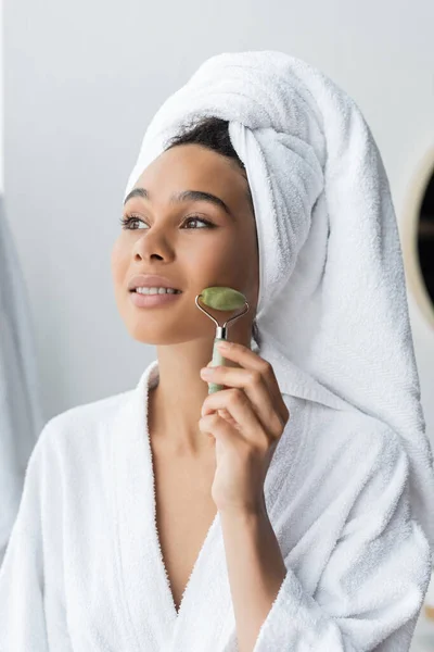 Smiling young african american woman in bathrobe and towel massaging face with jade roller — Stock Photo