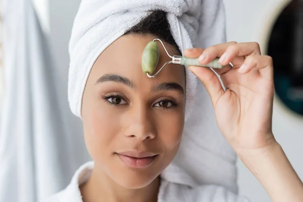 Close up of african american woman in white towel massaging forehead with jade roller in bathroom — Stock Photo