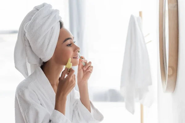 Vista lateral de mujer afroamericana en albornoz masajeando la cara con rodillo de jade y rascador facial en el baño - foto de stock