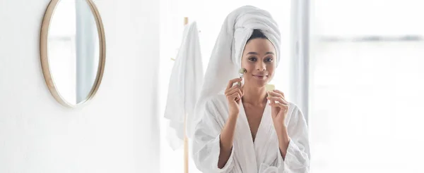 Happy african american woman in bathrobe massaging face with jade roller and face scraper, banner — Stock Photo