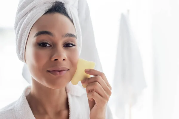 Jovem mulher afro-americana em toalha branca massageando rosto com raspador de rosto jade — Fotografia de Stock