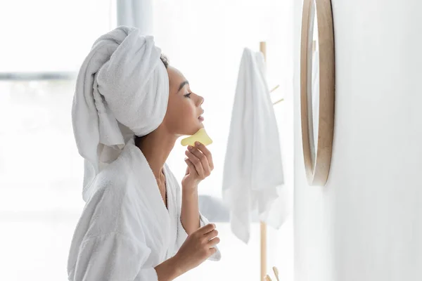 Side view of young african american woman in white towel scrapping face with jade face tool — Stock Photo