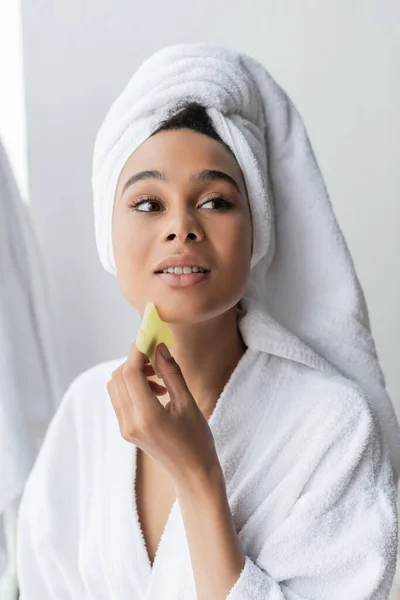 Mujer afroamericana bastante joven en toalla blanca masajeando la cara con raspador de jade - foto de stock