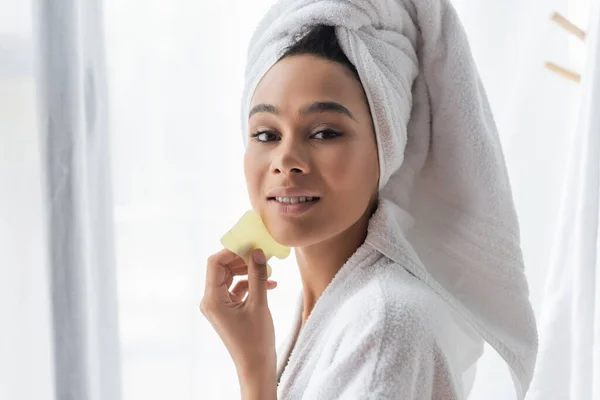 Mulher americana africana bonita em toalha branca segurando jade cara raspador perto do rosto — Fotografia de Stock