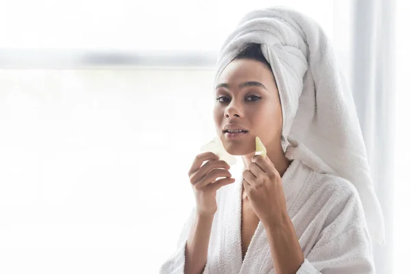 Giovane donna afroamericana in asciugamano bianco massaggiante viso con raschietti viso di giada — Foto stock