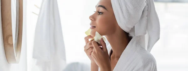 Vue latérale de la jeune femme afro-américaine qui gratte le visage avec des grattoirs en jade et qui regarde le miroir, la bannière — Photo de stock