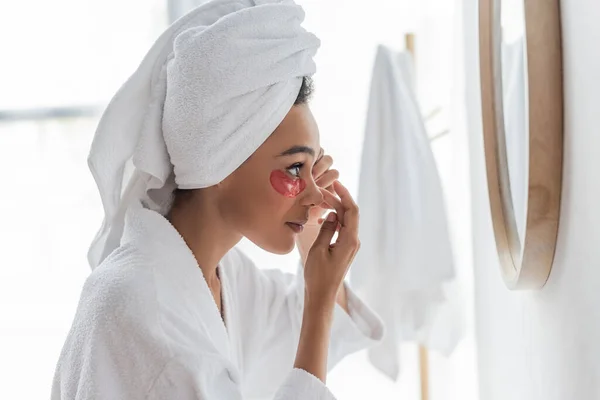 Side view of young african american woman applying eye patch while looking at mirror — Stock Photo