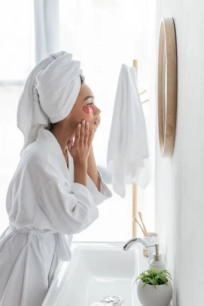 Vista laterale della donna afro-americana che applica benda sull'occhio mentre guarda lo specchio — Foto stock