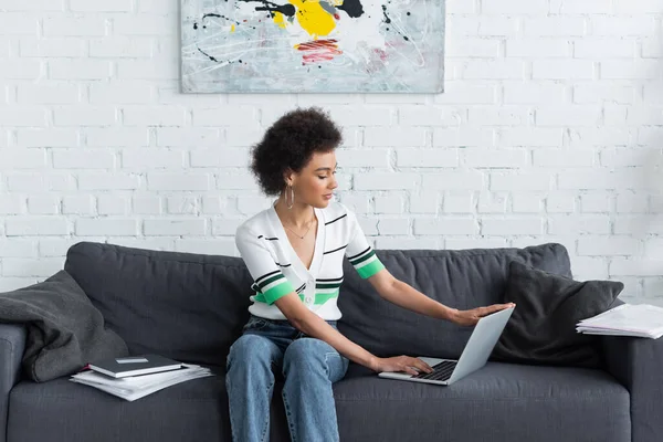 Encaracolado afro-americano mulher usando laptop perto de papéis e cartão de crédito no sofá — Fotografia de Stock