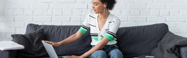 Curly african american woman using laptop near papers on sofa, banner — Stock Photo