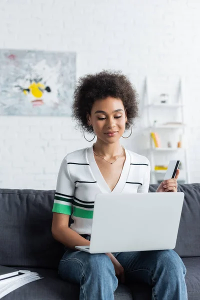 Mujer afroamericana rizada utilizando el ordenador portátil mientras sostiene la tarjeta de crédito en la sala de estar - foto de stock