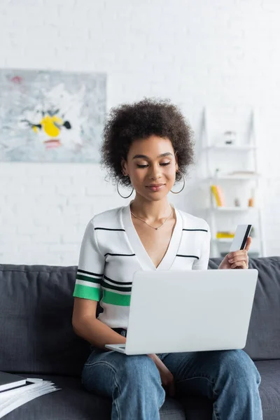 Frisé afro-américaine femme en utilisant un ordinateur portable tout en tenant la carte de crédit dans le salon — Photo de stock