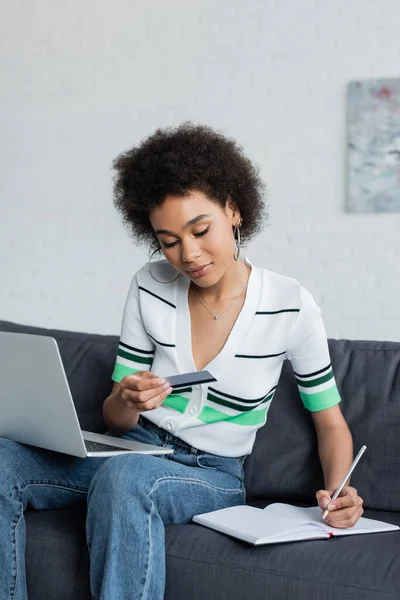 Curly african american woman with laptop holding credit card and writing in notebook in living room — Stock Photo
