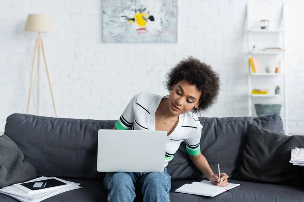 Lockige Afroamerikanerin schaut auf Laptop und schreibt in Notizbuch im Wohnzimmer — Stockfoto