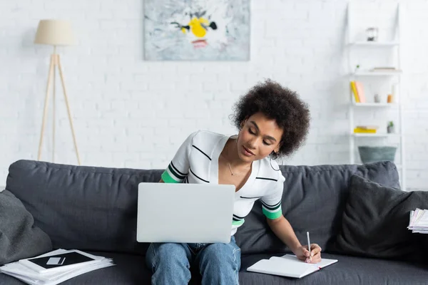 Mujer americana africana rizada mirando el ordenador portátil y la escritura en el cuaderno en la sala de estar - foto de stock