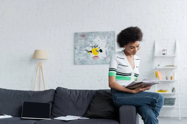 Femme afro-américaine bouclée regardant des documents près d'un ordinateur portable avec écran blanc sur le canapé — Photo de stock