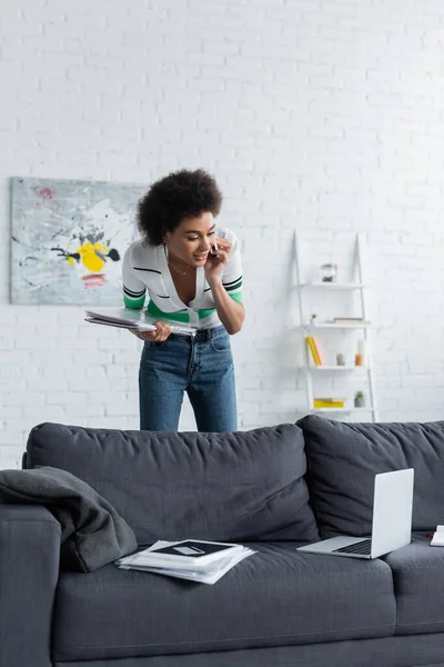 Lockige Afroamerikanerin schaut auf Laptop, während sie im Wohnzimmer mit dem Smartphone spricht — Stockfoto