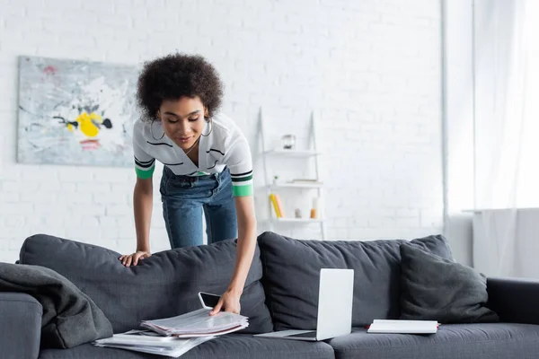 Lockige Afroamerikanerin greift nach Dokumenten, während sie ihr Smartphone in der Nähe des Laptops auf der Couch hält — Stockfoto