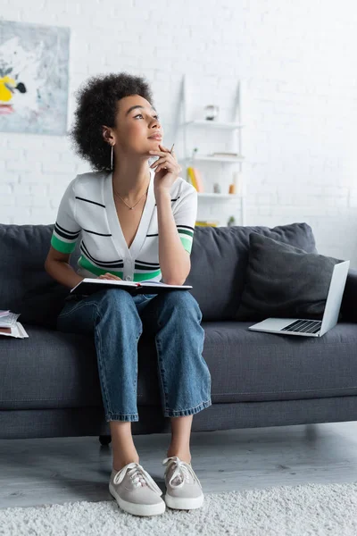 Nachdenkliche Afroamerikanerin sitzt mit Notizbuch neben Laptop auf Couch — Stockfoto