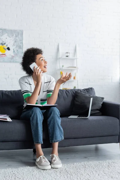 Счастливая африканская американка сидит с ноутбуком и разговаривает на смартфоне возле ноутбука на диване — стоковое фото