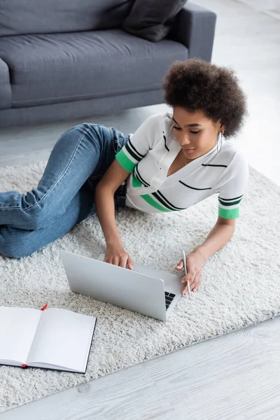 African american freelancer using laptop while lying on carpet — Stock Photo