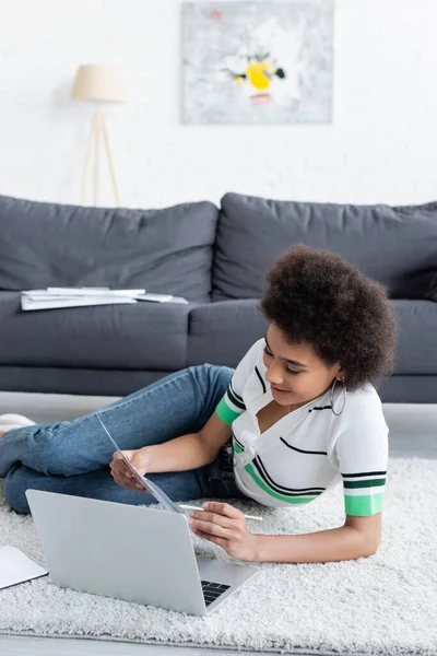 Cheerful african american freelancer looking at document near laptop while lying on carpet — Stock Photo