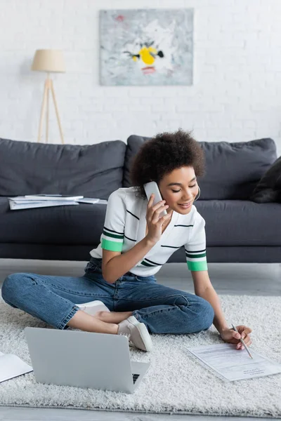 Souriant afro-américain pigiste parler sur smartphone près d'un ordinateur portable tout en étant assis sur le tapis — Photo de stock