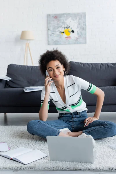 Cheerful african american freelancer talking on smartphone near laptop while sitting on carpet — Stock Photo