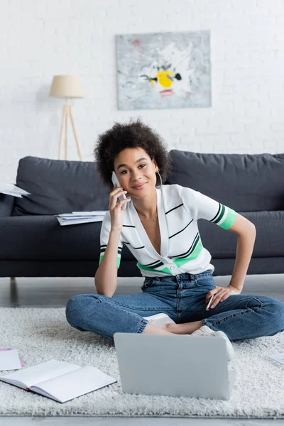 Allegro freelance afroamericano che parla su smartphone vicino al computer portatile mentre è seduto su un tappeto — Foto stock