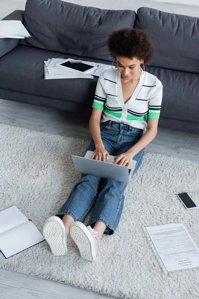 Vista ad alto angolo di freelance americano africano utilizzando il computer portatile e seduto su tappeto — Foto stock