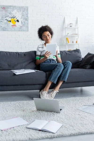Mujer afroamericana usando tableta digital mientras está sentada en el sofá - foto de stock