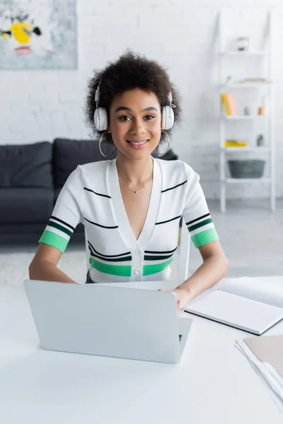 Femme afro-américaine gaie dans des écouteurs sans fil près d'un ordinateur portable sur le bureau — Photo de stock