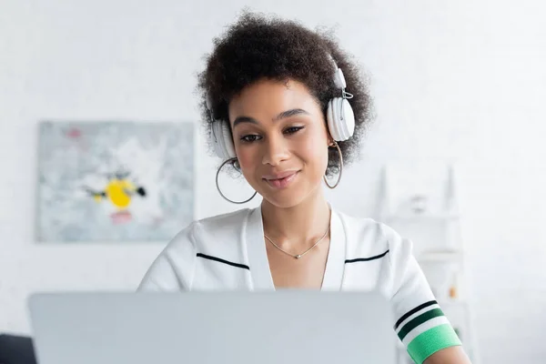 Pleased african american woman in wireless headphones looking at blurred laptop — Stock Photo