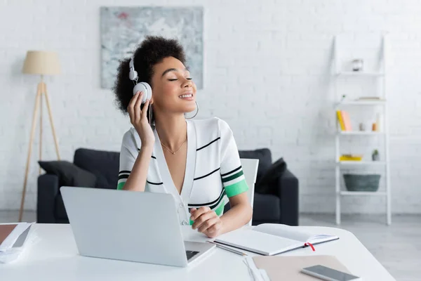 Mujer afroamericana feliz en auriculares inalámbricos escuchando música cerca de gadgets - foto de stock