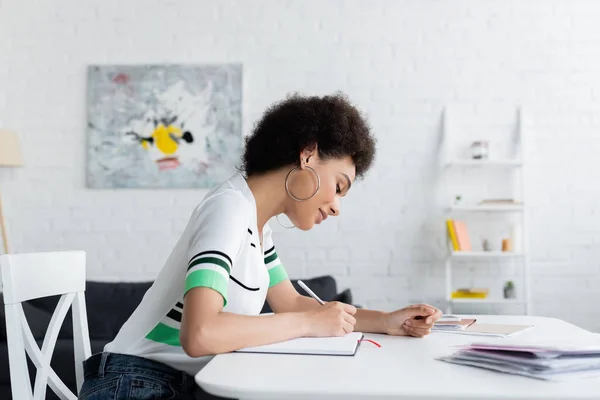 Vista laterale della donna afro-americana che scrive sul taccuino vicino ai giornali a casa — Foto stock