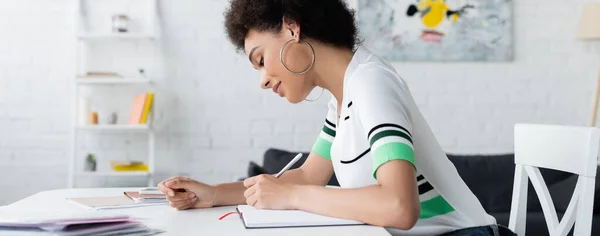 Vista lateral de una mujer afroamericana escribiendo en un cuaderno cerca de papeles en casa, pancarta - foto de stock