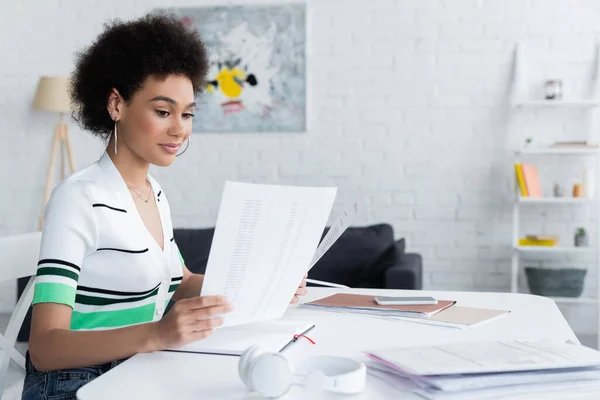 Jeune femme afro-américaine tenant des papiers près des écouteurs et smartphone à la maison — Photo de stock