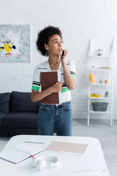 Africano americano mulher falando no smartphone e segurando pasta de papel em casa — Fotografia de Stock