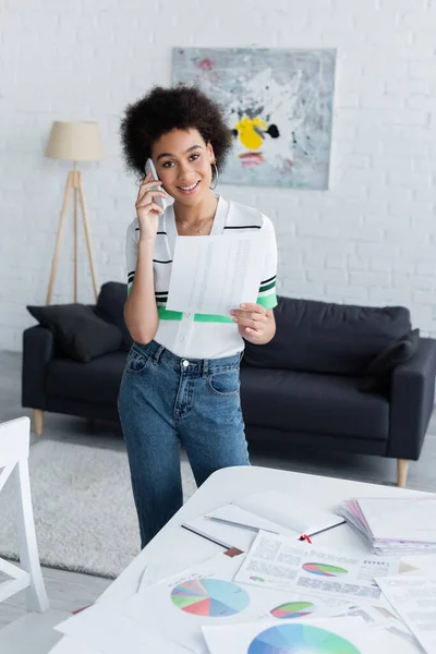 Positive african american woman talking on smartphone near papers with charts at home — Stock Photo