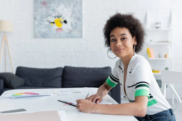 Lächelnde Afroamerikanerin blickt in die Kamera neben Notizbuch und Dokumenten zu Hause — Stockfoto