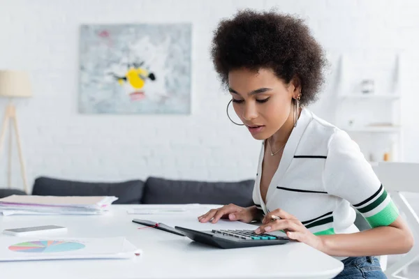 Africano americano mulher usando calculadora perto de gráficos na mesa em casa — Fotografia de Stock